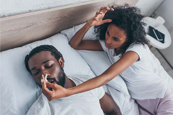 Treatment for sleep apnea is here. Sleep Better Columbus Ohio can help you get the best sleep possible so that you can enjoy your life again! Quit suffering and visit us today. Image of couple laying in bed. Man is snoring and woman trying to put a clamp on his nose.